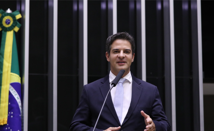 Deputado discursa no plenário da Câmara com a bandeira do Brasil ao fundo.