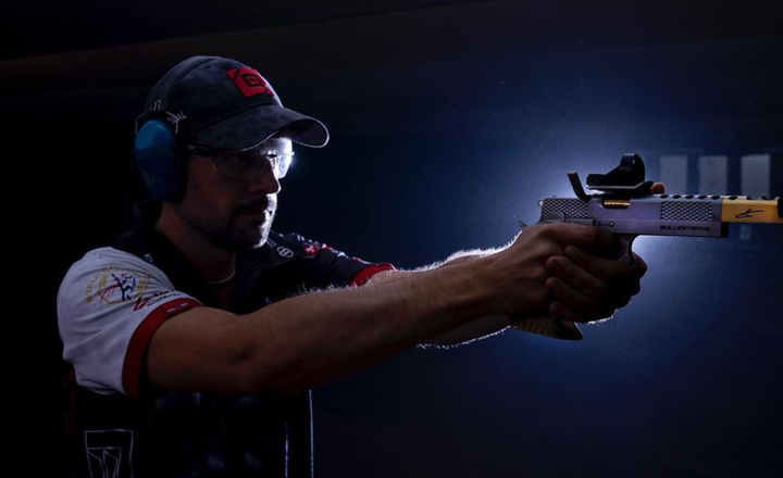 Jorge Ballesteros atira com uma pistola equipada com mira em ambiente escuro.