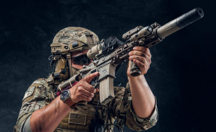 Atirador com roupa tática usa um fuzil equipado com mira e silenciador.