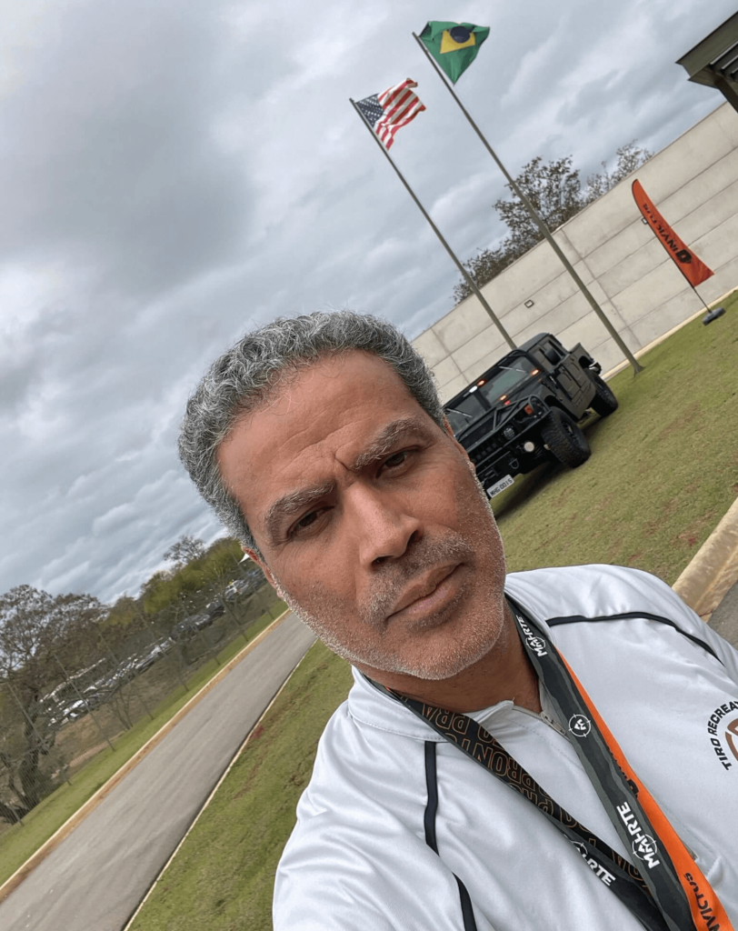Demetrius Oliveira tira selfie em campo de tiro com bandeiras ao fundo.
