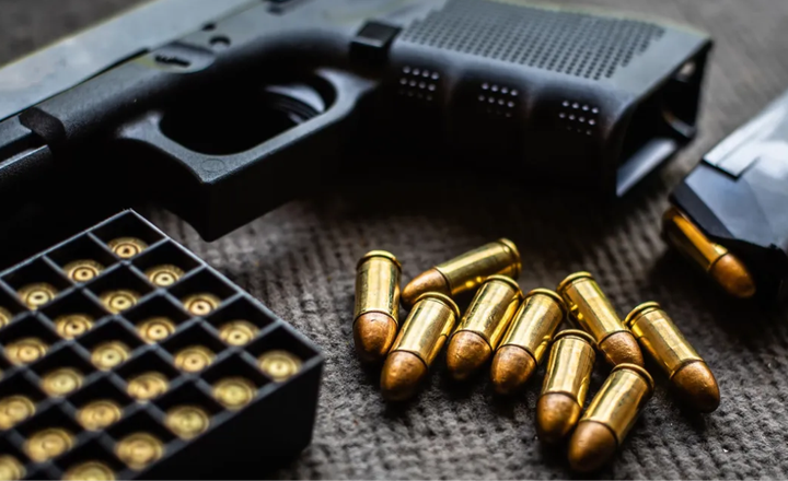 Pistola, caixa de munição e cartuchos espalhados sobre uma mesa escura.