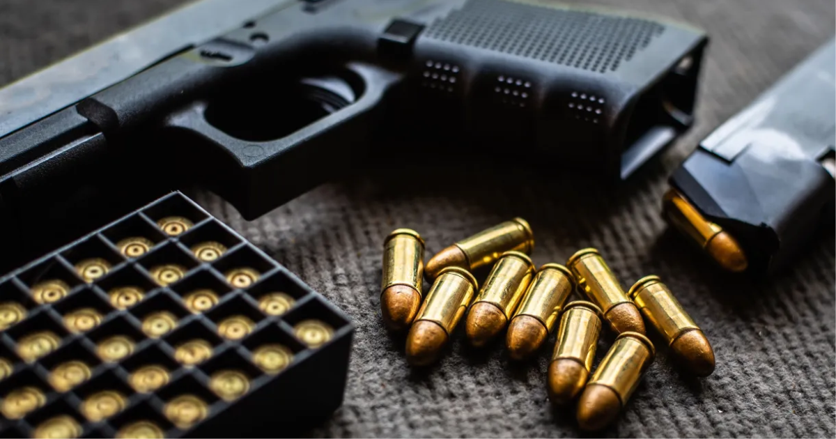 Pistola, caixa de munição e cartuchos espalhados sobre uma mesa escura.