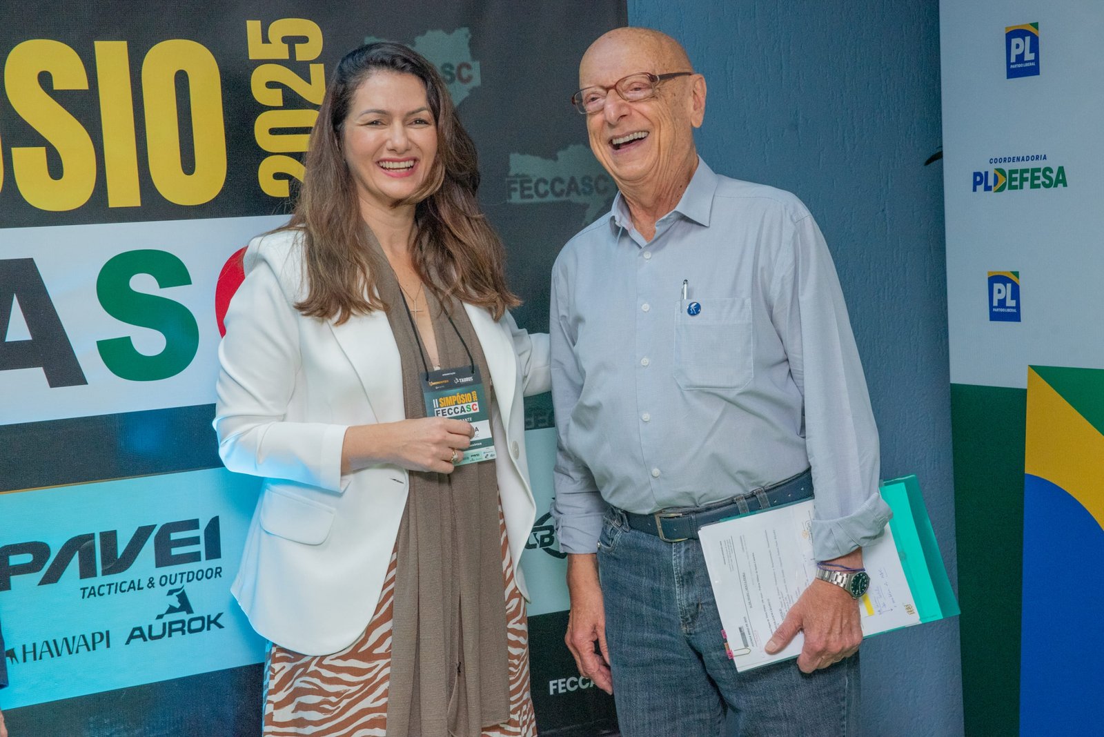 Mulher e homem idoso sorriem diante do painel do Simpósio FECCASC.
