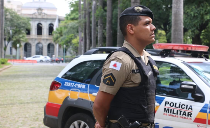 Policial militar de Minas Gerais diante de viatura e prédio público.