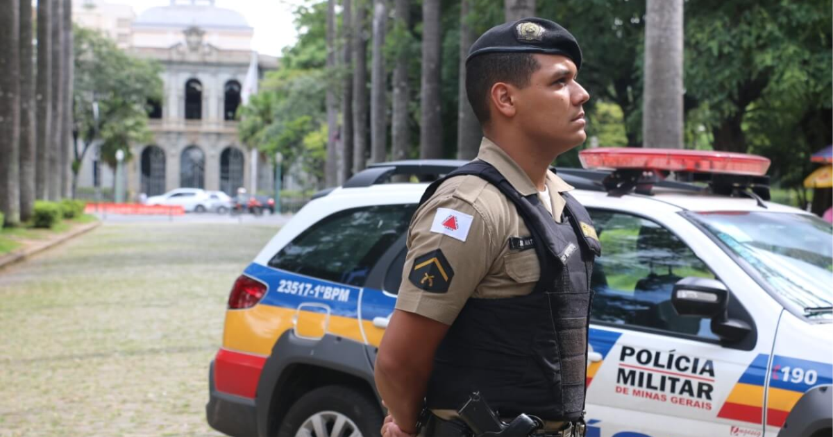 Policial militar de Minas Gerais diante de viatura e prédio público.