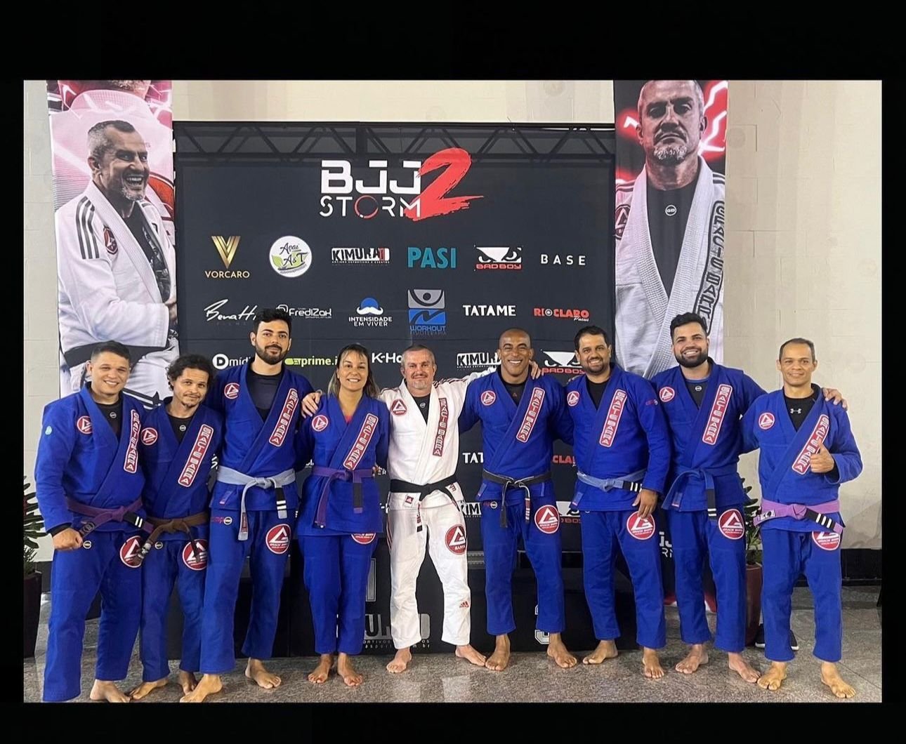 Equipe de jiu-jítsu posa reunida diante de painel de competição.