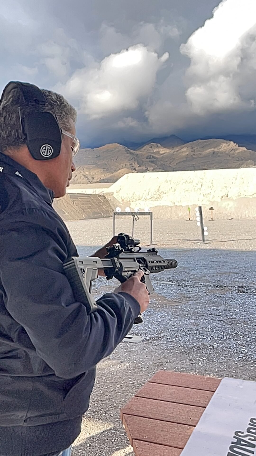 Atirador usa arma longa em campo de tiro cercado por montanhas.