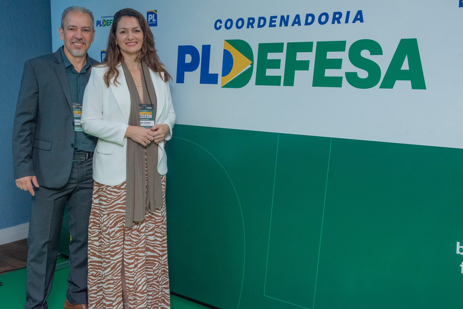 Homem e mulher posam diante do painel da Coordenadoria PL Defesa.