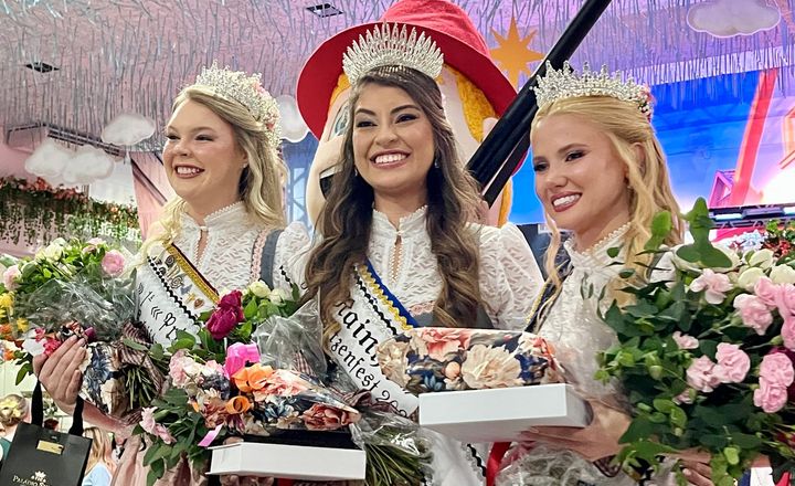 Três rainhas da Schützenfest posam com coroas, flores e um rifle cerimonial.