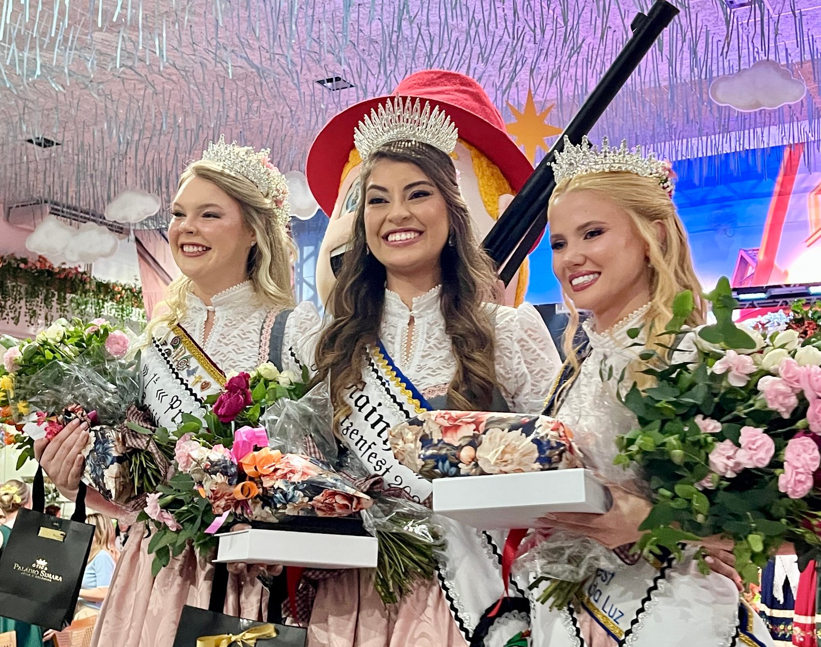 Três rainhas da Schützenfest posam com coroas, flores e um rifle cerimonial.