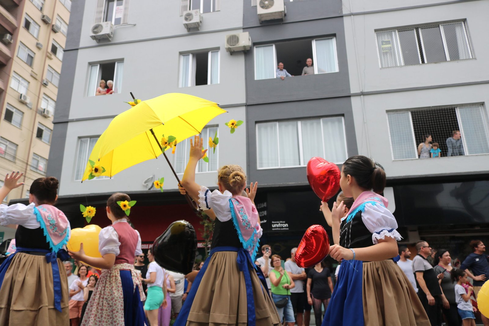 Participantes em trajes típicos dançam com guarda-chuvas coloridos durante desfile.