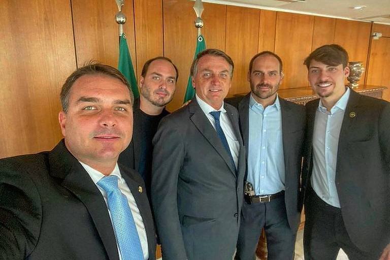 Cinco homens posam juntos em uma sala com painéis de madeira e bandeiras brasileiras.