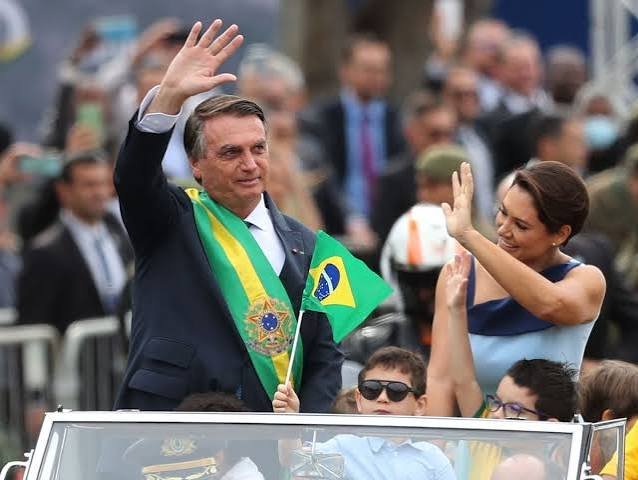 Homem com a faixa presidencial acena de um carro aberto, acompanhado por uma mulher e uma criança.
