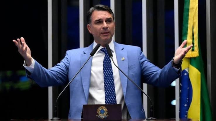 Homem de terno azul discursa com os braços abertos em uma tribuna, diante de uma bandeira brasileira.