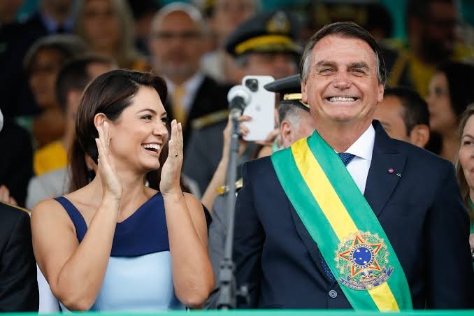 Homem com a faixa presidencial sorri ao lado de uma mulher durante uma cerimônia com público ao fundo.