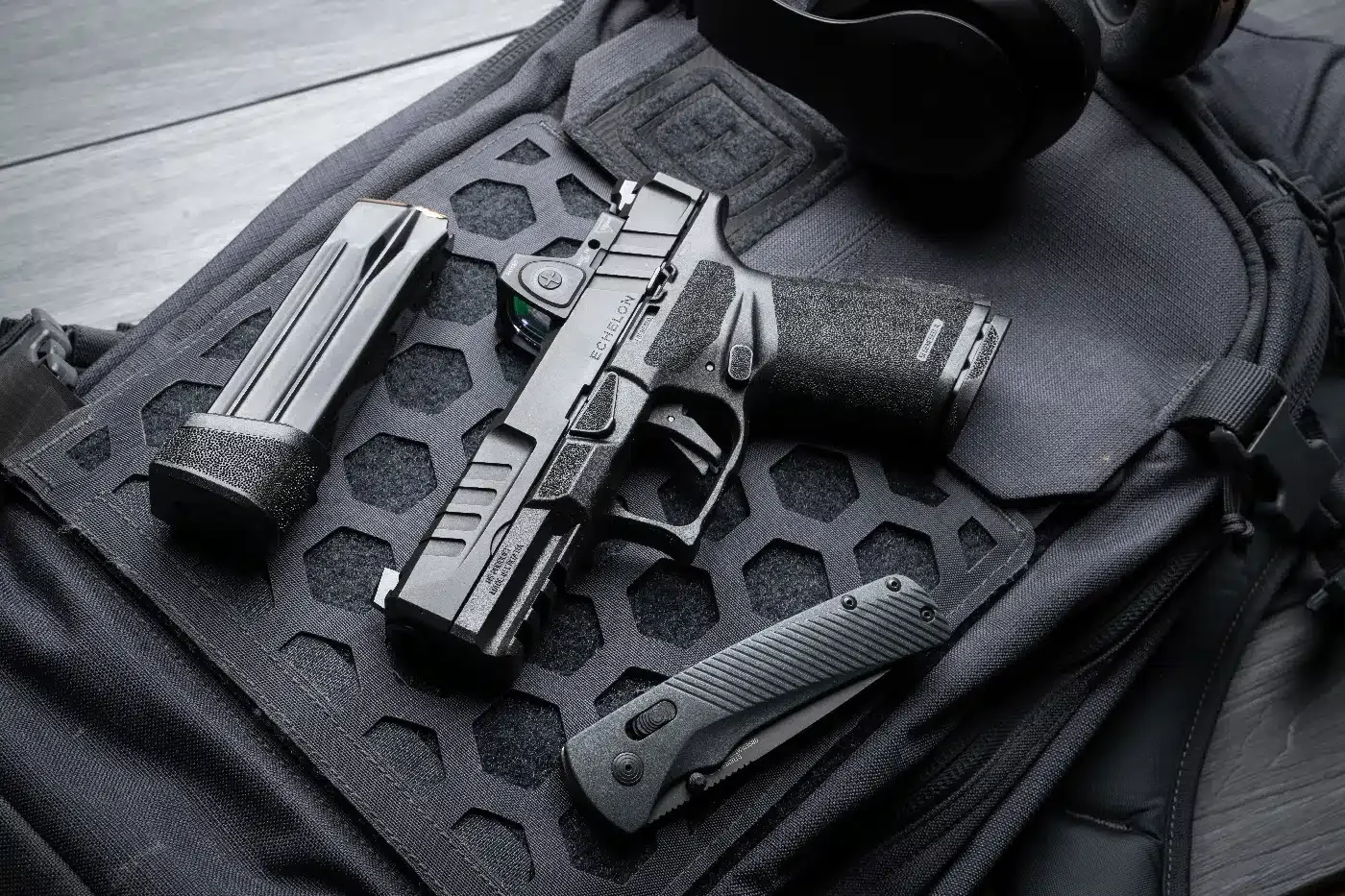 Pistola, carregador e canivete apoiados sobre uma bolsa tática preta.