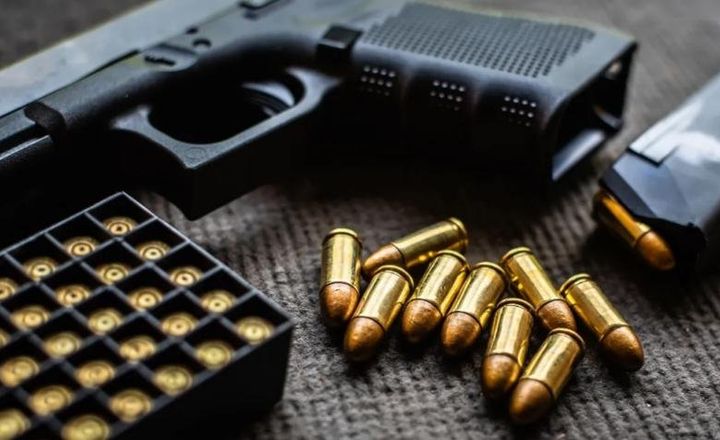 Pistola, caixa de munição e cartuchos espalhados sobre uma mesa escura.
