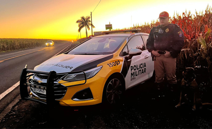 Policial militar ao lado de viatura em rodovia durante o pôr do sol.