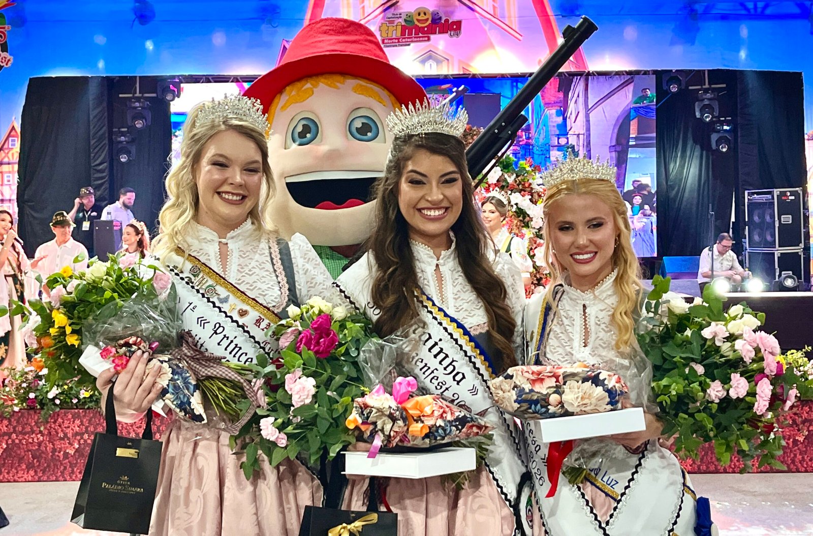 Rainhas da Schützenfest posam com coroas, faixas, flores e rifle cerimonial.