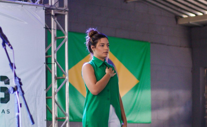 Mulher fala ao microfone em palco diante de uma bandeira do Brasil.