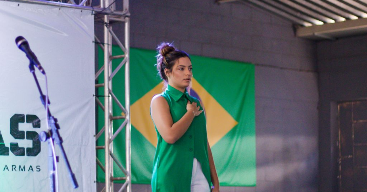 Mulher fala ao microfone em palco diante de uma bandeira do Brasil.