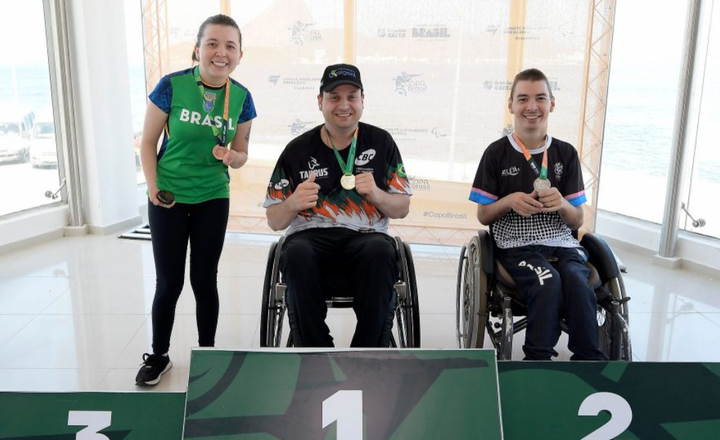 Três atletas do tiro paralímpico exibem medalhas em um pódio.