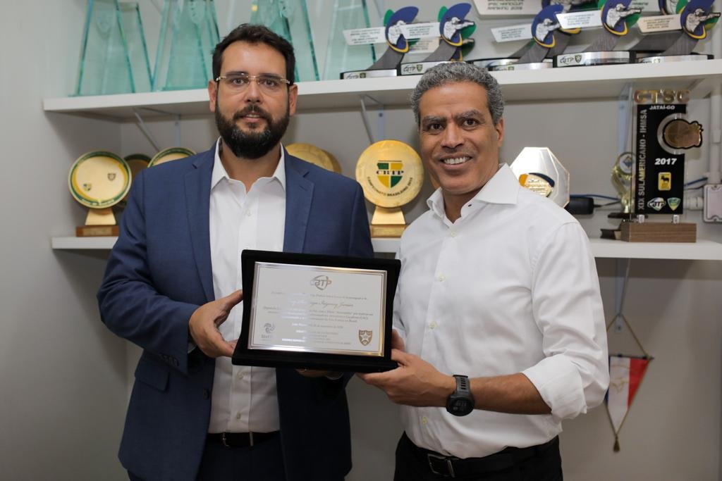 Demetrius Oliveira e outro homem exibem certificado diante de troféus.