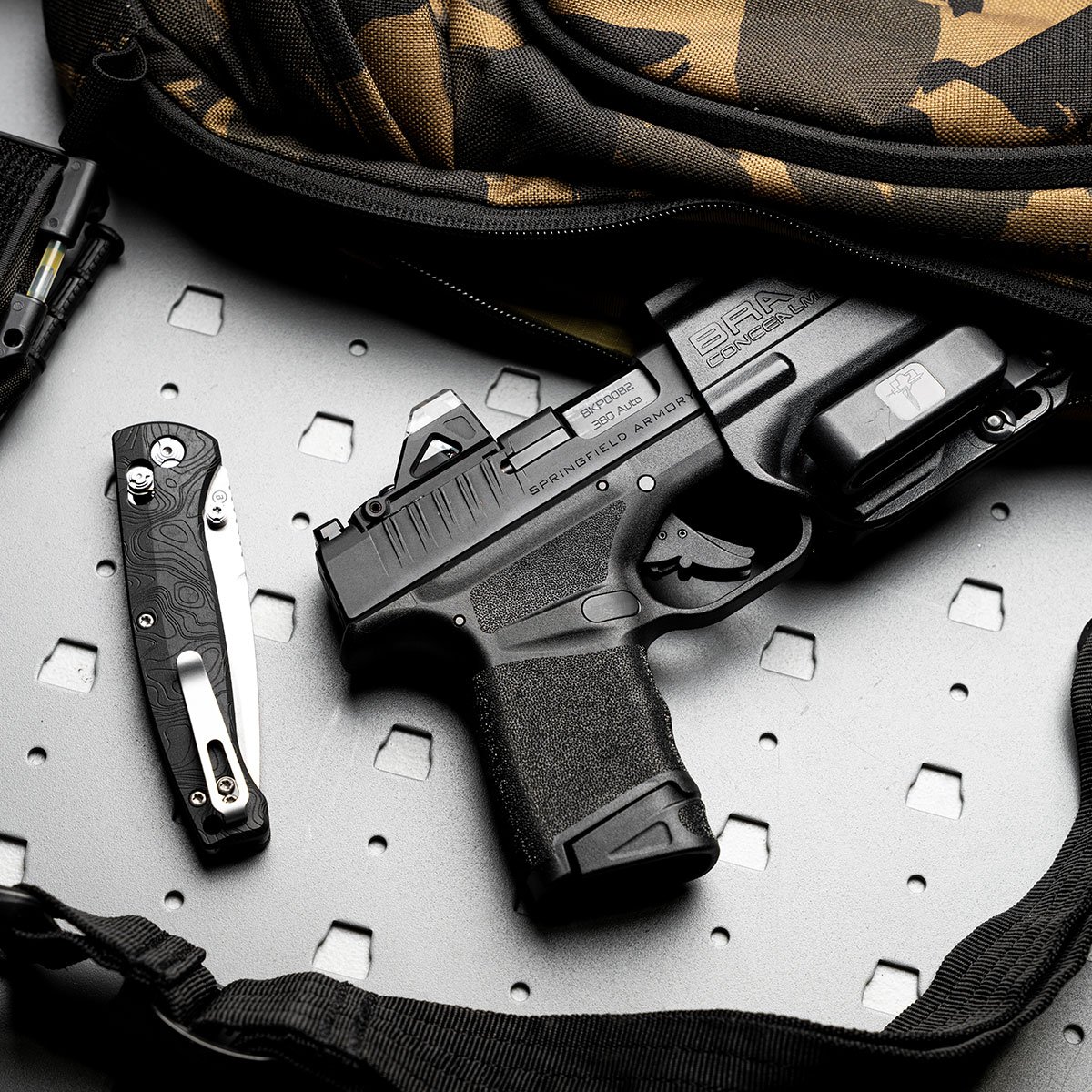 Pistola Springfield Armory Hellcat e canivete sobre superfície metálica.