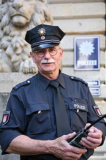 Policial alemão uniformizado segura um rádio diante de uma delegacia.