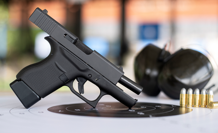 Pistola preta, óculos de proteção e cartuchos sobre um alvo de papel.