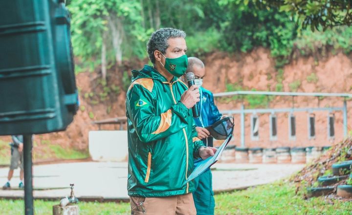 Demetrius Oliveira fala ao microfone em evento de tiro ao ar livre.